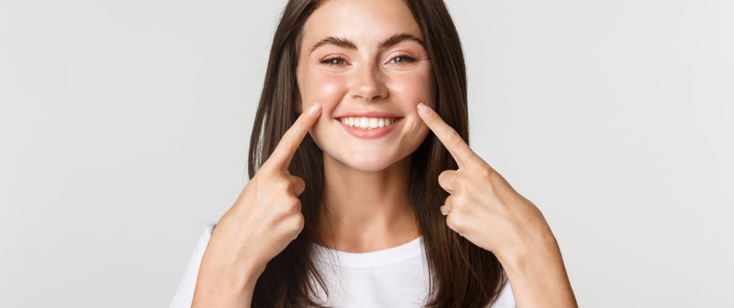 Close-up of attractive smiling young girl pointing fingers at face, poking cheeks, white background